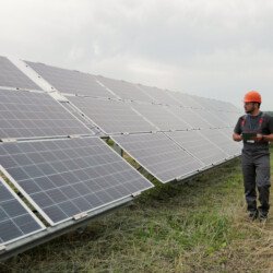Male Engineer In Hard Hat Walks On Renewable Energy Station Holding Digital Tablet And SBI  Scaled